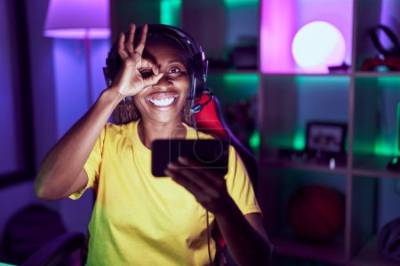 Foto de Mujer afroamericana jugando videojuegos con smartphone sonriendo feliz haciendo ok signo con la mano en el ojo mirando a través de los dedos - Imagen libre de derechos