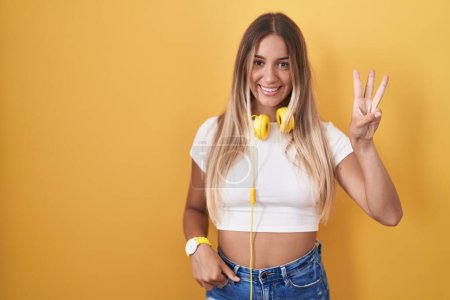 Photo for Young blonde woman standing over yellow background wearing headphones showing and pointing up with fingers number three while smiling confident and happy. - Royalty Free Image