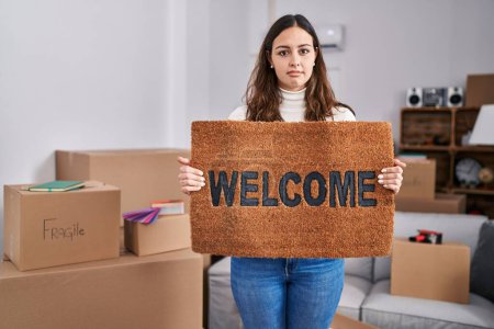 Photo for Young hispanic woman holding welcome doormat at new home relaxed with serious expression on face. simple and natural looking at the camera. - Royalty Free Image