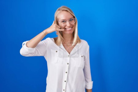 Téléchargez les photos : Jeune femme caucasienne debout sur fond bleu souriant faire geste de téléphone avec la main et les doigts comme parler au téléphone. communiquer les concepts. - en image libre de droit