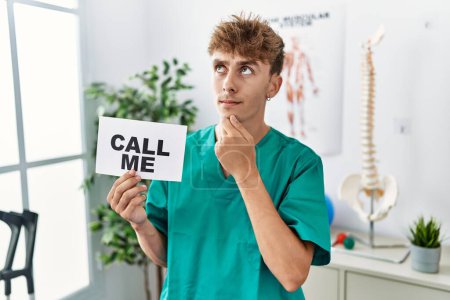 Photo for Young caucasian physio man holding call me banner at the clinic serious face thinking about question with hand on chin, thoughtful about confusing idea - Royalty Free Image