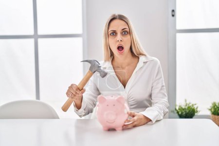 Foto de Young blonde woman holding piggy bank and hammer in shock face, looking skeptical and sarcastic, surprised with open mouth - Imagen libre de derechos