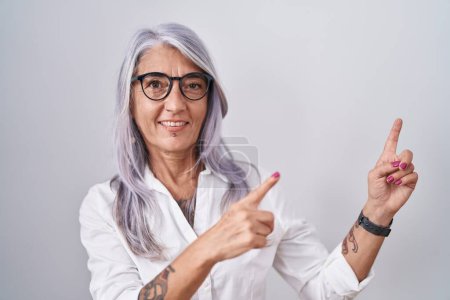 Téléchargez les photos : Femme d'âge moyen avec des tatouages portant des lunettes debout sur fond blanc souriant et regardant la caméra pointant avec deux mains et doigts sur le côté. - en image libre de droit