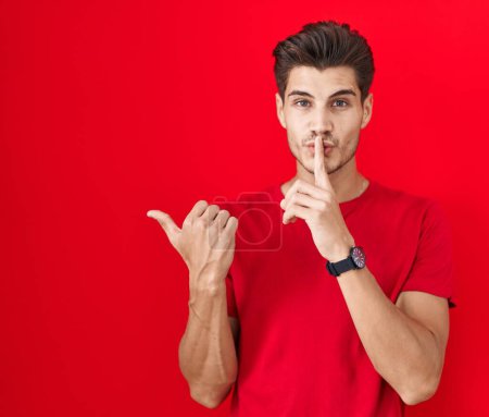 Téléchargez les photos : Young hispanic man standing over red background asking to be quiet with finger on lips pointing with hand to the side. silence and secret concept. - en image libre de droit