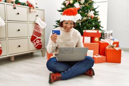 Foto de Mujer hispana de mediana edad con sombrero de navidad sosteniendo regalo y tarjeta de crédito con un aspecto positivo y feliz de pie y sonriendo con una sonrisa confiada mostrando los dientes - Imagen libre de derechos