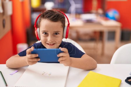 Photo for Adorable hispanic toddler student using smartphone and headphones studying at classroom - Royalty Free Image