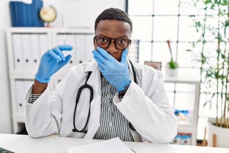 Téléchargez les photos : Jeune homme médecin africain tenant seringue à l'hôpital choqué couvrant la bouche avec les mains pour erreur. concept secret. - en image libre de droit