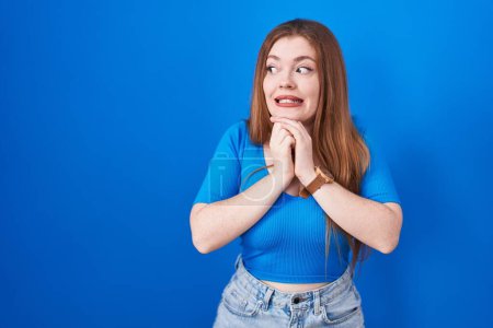 Foto de Redhead woman standing over blue background laughing nervous and excited with hands on chin looking to the side - Imagen libre de derechos