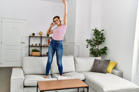 Téléchargez les photos : Jeune femme écoutant de la musique et dansant debout sur le canapé à la maison - en image libre de droit