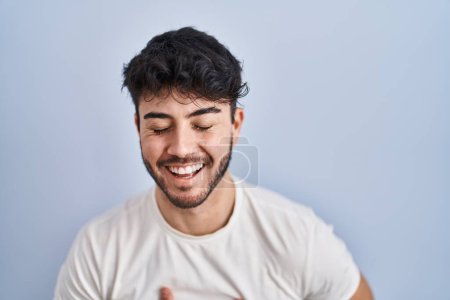 Foto de Hombre hispano con barba de pie sobre fondo blanco sonriendo y riendo en voz alta porque divertido chiste loco con las manos en el cuerpo. - Imagen libre de derechos