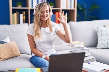 Foto de Mujer rubia joven estudiando el uso de ordenador portátil en casa renunciando a decir hola feliz y sonriente, gesto de bienvenida amistoso - Imagen libre de derechos