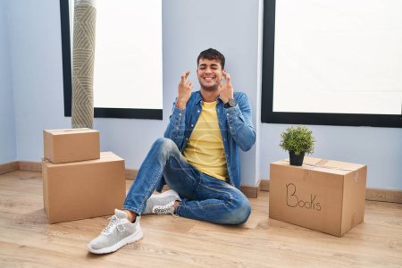Foto de Young hispanic man sitting on the floor at new home gesturing finger crossed smiling with hope and eyes closed. luck and superstitious concept. - Imagen libre de derechos