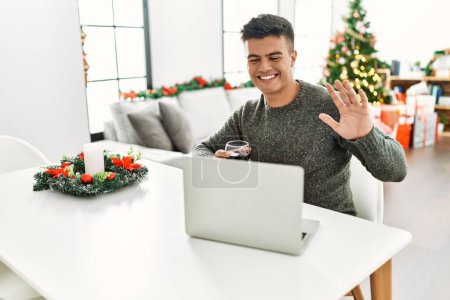 Téléchargez les photos : Jeune homme hispanique buvant du vin ayant appel vidéo assis près de l'arbre de Noël à la maison - en image libre de droit