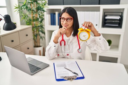 Photo for Young hispanic doctor woman holding alarm clock at the clinic serious face thinking about question with hand on chin, thoughtful about confusing idea - Royalty Free Image