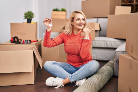 Photo for Blonde woman holding keys of new home sitting on the floor pointing thumb up to the side smiling happy with open mouth - Royalty Free Image