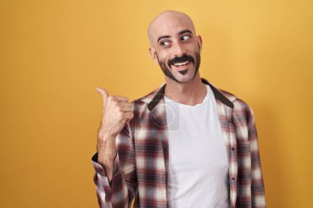 Téléchargez les photos : Homme hispanique avec la barbe debout sur fond jaune souriant avec le visage heureux regardant et pointant vers le côté avec le pouce vers le haut. - en image libre de droit
