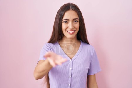 Foto de Joven mujer hispana con el pelo largo de pie sobre fondo rosa sonriente alegre ofreciendo la palma de la mano dando asistencia y aceptación. - Imagen libre de derechos