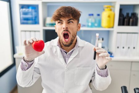 Foto de Arab man with beard working at scientist laboratory holding blood samples angry and mad screaming frustrated and furious, shouting with anger. rage and aggressive concept. - Imagen libre de derechos