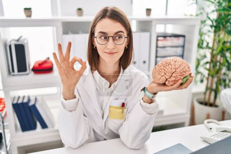 Photo for Young caucasian doctor woman holding brain as mental health concept doing ok sign with fingers, smiling friendly gesturing excellent symbol - Royalty Free Image
