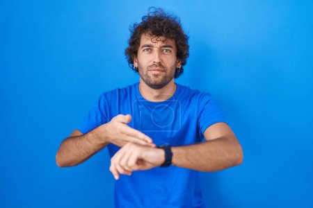 Téléchargez les photos : Hispanic young man standing over blue background in hurry pointing to watch time, impatience, upset and angry for deadline delay - en image libre de droit