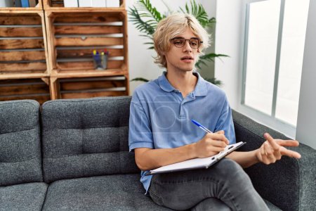 Téléchargez les photos : Jeune homme blond psychologue écrit sur document parlant à la clinique de psychologie - en image libre de droit