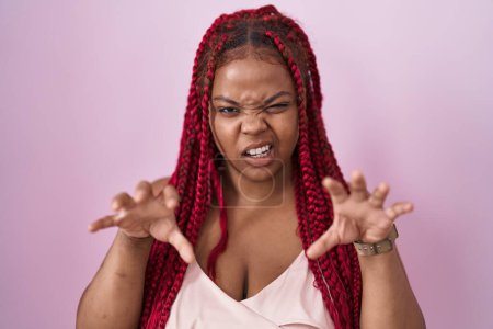 Téléchargez les photos : Femme afro-américaine avec des cheveux tressés debout sur fond rose souriant drôle faisant griffe geste comme chat, expression agressive et sexy - en image libre de droit