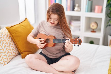 Téléchargez les photos : Jeune rousse femme jouant ukulele assis sur le lit dans la chambre - en image libre de droit