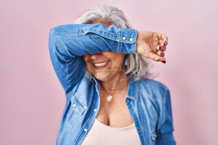 Téléchargez les photos : Femme d'âge moyen avec les cheveux gris debout sur fond rose couvrant les yeux avec le bras souriant gai et drôle. concept aveugle. - en image libre de droit