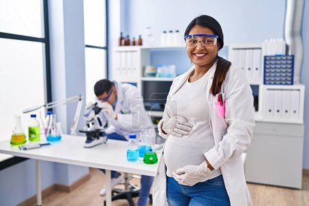 Téléchargez les photos : Young hispanic woman expecting a baby working at scientist laboratory looking positive and happy standing and smiling with a confident smile showing teeth - en image libre de droit