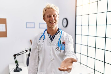 Photo for Young blond man wearing doctor uniform and stethoscope at clinic smiling cheerful offering palm hand giving assistance and acceptance. - Royalty Free Image