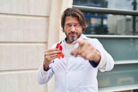Téléchargez les photos : Beau médecin d'âge moyen homme tenant soutien ruban rouge pointant avec le doigt à la caméra et à vous, geste confiant regardant sérieux - en image libre de droit