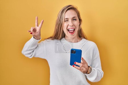 Foto de Young blonde woman using smartphone typing message smiling with happy face winking at the camera doing victory sign with fingers. number two. - Imagen libre de derechos