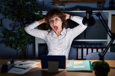 Téléchargez les photos : Middle age woman working at the office at night crazy and scared with hands on head, afraid and surprised of shock with open mouth - en image libre de droit