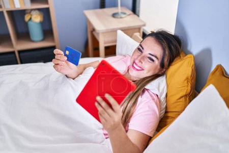 Foto de Joven hermosa mujer hispana usando touchpad y tarjeta de crédito acostada en la cama en el dormitorio - Imagen libre de derechos