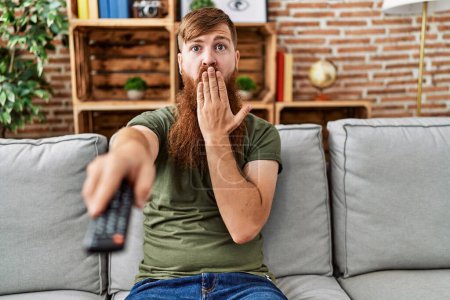 Téléchargez les photos : Homme rousse à longue barbe tenant télécommande de télévision couvrant la bouche avec la main, choqué et effrayé par erreur. expression surprise - en image libre de droit