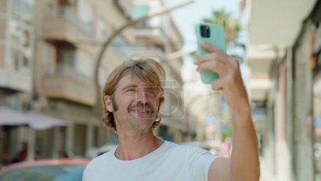 Foto de Joven hombre rubio sonriendo confiado haciendo selfie por el teléfono inteligente en la calle - Imagen libre de derechos