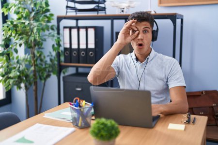 Photo for Young hispanic man working at the office wearing headphones doing ok gesture shocked with surprised face, eye looking through fingers. unbelieving expression. - Royalty Free Image