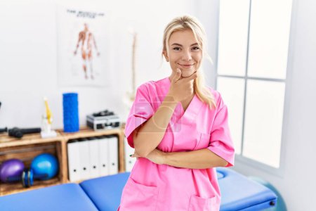 Téléchargez les photos : Young caucasian woman working at pain recovery clinic looking confident at the camera with smile with crossed arms and hand raised on chin. thinking positive. - en image libre de droit