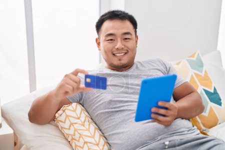 Téléchargez les photos : Young chinese man using touchpad and credit card sitting on bed at bedroom - en image libre de droit