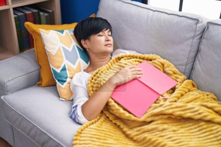 Photo for Middle age chinese woman holding book lying on sofa sleeping at home - Royalty Free Image