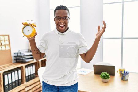 Téléchargez les photos : Jeune homme africain tenant un réveil au bureau célébrant la victoire avec un sourire heureux et l'expression gagnante avec les mains levées - en image libre de droit