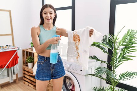 Photo for Young hispanic woman cleaning dirty white t shirt with stain at laundry room winking looking at the camera with sexy expression, cheerful and happy face. - Royalty Free Image