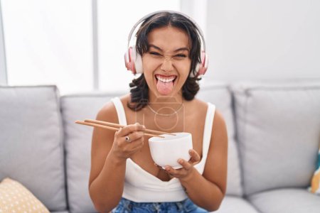 Foto de Young hispanic woman eating asian food using chopsticks sticking tongue out happy with funny expression. - Imagen libre de derechos