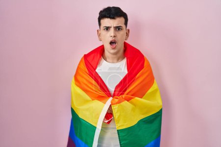 Téléchargez les photos : Non binary person holding rainbow lgbtq flag scared and amazed with open mouth for surprise, disbelief face - en image libre de droit