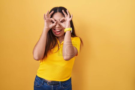 Photo for Young arab woman standing over yellow background doing ok gesture like binoculars sticking tongue out, eyes looking through fingers. crazy expression. - Royalty Free Image