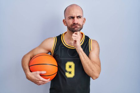 Foto de Joven calvo con barba llevando uniforme de baloncesto sosteniendo la pelota pensando concentrado en la duda con el dedo en la barbilla y mirando hacia arriba preguntándose - Imagen libre de derechos