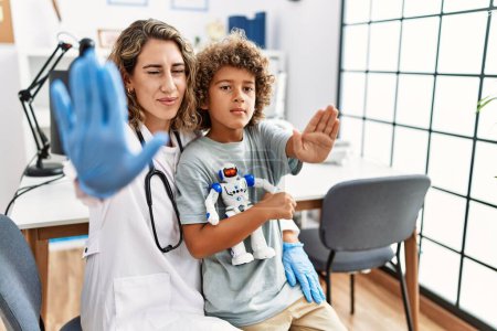 Foto de Young kid at pediatrician clinic holding teddy bear with open hand doing stop sign with serious and confident expression, defense gesture - Imagen libre de derechos
