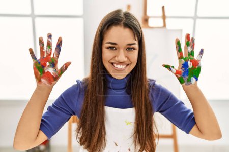Foto de Joven latina sonriendo confiada mostrando las manos pintadas de palma en el estudio de arte - Imagen libre de derechos