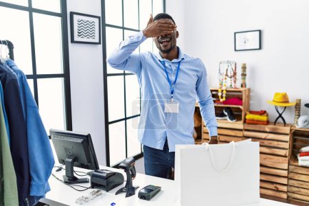 Foto de Joven hombre africano que trabaja como gerente en boutique minorista sonriendo y riendo con la mano en la cara cubriendo los ojos para sorpresa. concepto ciego. - Imagen libre de derechos