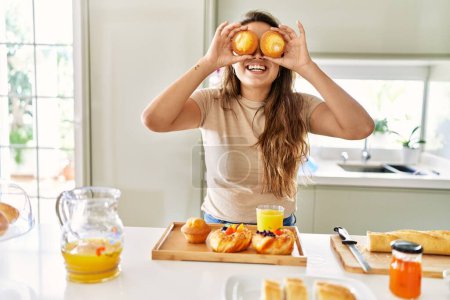 Téléchargez les photos : Jeune belle femme hispanique préparant le petit déjeuner tenant des cupcakes sur les yeux à la cuisine - en image libre de droit